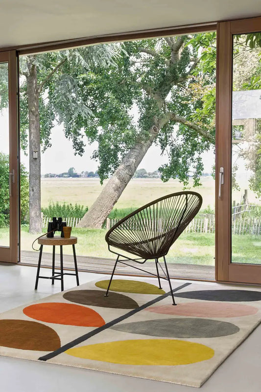 colorful leaf design hand tufted rug in a room with Modern chair, large windows showing greenery outside.
