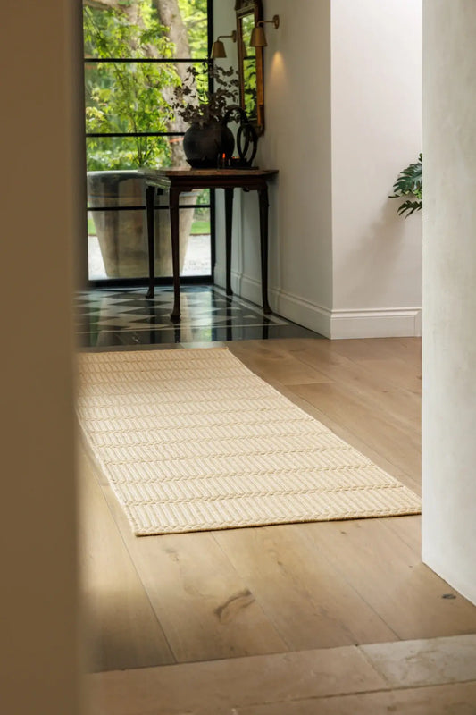Beige hand woven wool runner on a wooden floor with a glass door and plants in the background